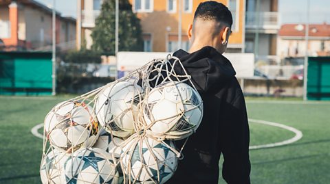 Football player carrying a net of footballs on pitch.
