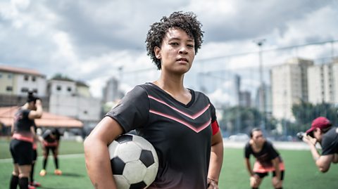 Female football player holding a football under her arm on a pitch.