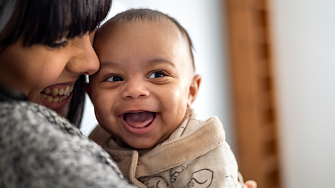 A very cheerful baby beams at something off camera, while their mum cuddles them, grinning.
