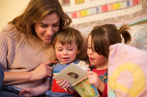 A mother enjoying a picture book on the sofa with her two young children.The older chid, a girl, seems to be showing the book to her younger brother.