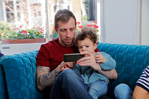 A father and toddler-aged daughter sit on the sofa together, watching something on his phone. 