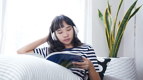 A teenage girl sits on a sofa, reading a book and listening to music through headphones.