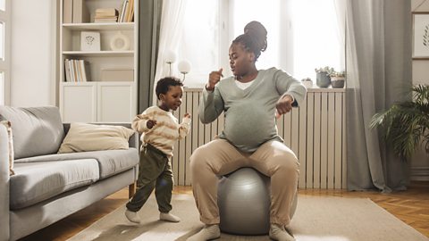 A mum bounces on an exercise ball in her living room with her son marching around her.