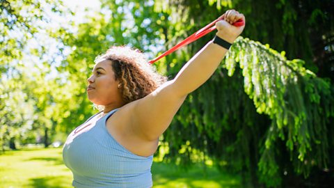 A woman in exercise gear pulls a resistance band behind her head.