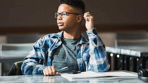 A teenager sits in a library with a notebook and headphones, building critical thinking skills