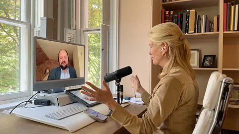 BBC BBC Special Correspondent Katty Kay speaks to author Ross Douthat on a video call. Kay is shown from behind with white earbuds and a black microphone and she wears a tan shirt. Douthat is on her monitor and can be seen wearing a dark blue jacket and black headphones (Credit: BBC)