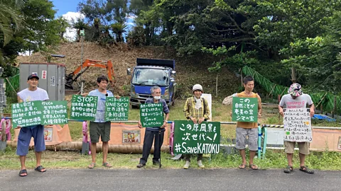 katoku.org A line of people stand with signs largely in Japanese. Some English words appear – "STOP", "UNESCO" and #Save Katoku" (Credit: katoku.org)