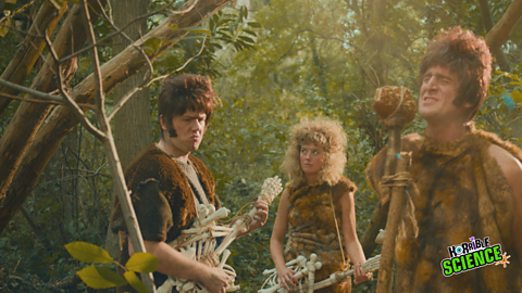 Still photograph of two men and a women dressed as pre-historic singing and playing guitars made of bones.