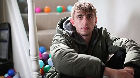 A young man sits at the bottom of an indoor staircase, looking seriously at the camera. Beside him and on the stairs are various coloured plastic balls.