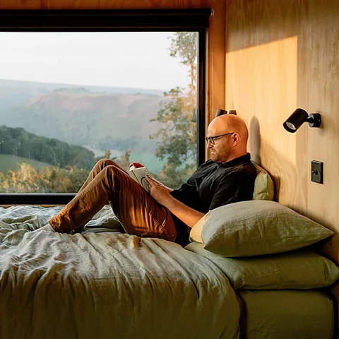 Adam Firman A man sits on a bed reading a book in a cabin which has views out to green hills (Credit: Adam Firman)