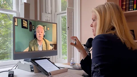 BBC BBC Special Correspondent Katty Kay is shown speaking to Edward Luce via videoconference. Luce can be seen on Kay's monitor wearing a tan jacket and over-ear headphones. Kay is seen from behind wearing a dark navy jacket, speaking into a black tabletop microphone (Credit: BBC)