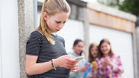 A young girl looking at her screen while a group of other young girls are behind her laughing at her.