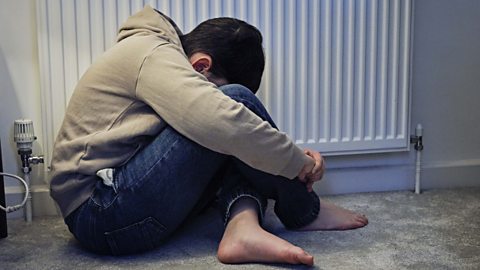 Young boy sitting beside a radiator hunched over cowering with his head on his knees.