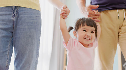 A young girl grins while holding her parents' hands.