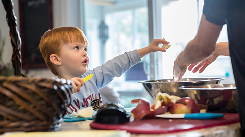 A young boy holds a child-safe knife, helping one of his parents with the cooking.