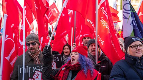 A group of workers are taking part in a protest carrying a number of red NIPSA (Trade Union) flags and one Unison (Trade Union) flag.