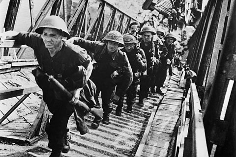 A line of soldiers walk across a stepped bridge on D-Day 1944