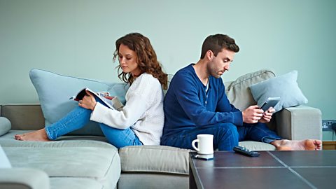 A woman and man sitting on a sofa back to back, the woman is reading a magazine and the man is holding a tablet.