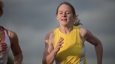 A girl running in a yellow and white athletic vest with another girl runner off to the side of her at the edge of the frame.