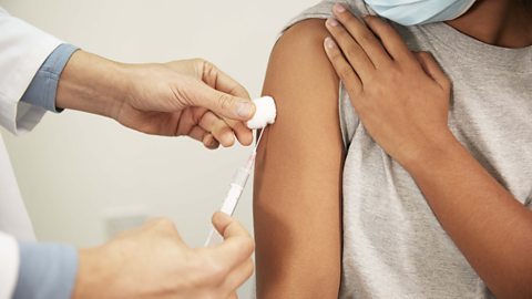 Doctor administering the Covid-19 vaccine in a syringe to a girl's right arm with a cotton wool pad beside the puncture wound.