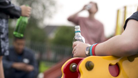 Some teenagers drinking alcohol in a play park, one is swigging from a bottle in the background and two are holding bottles in the forground.