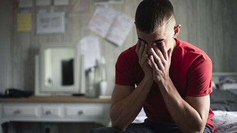 Young boy sitting in his bedroom with his head in his hands, stressed, with pages of schoolwork posted on his wall behind him.