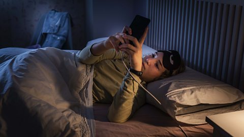 A girl is lying in bed with a sleep mask on her pulled up and on her phone in the middle of the night.