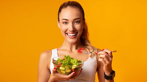 Girl in workout clothes eating a bowl of salad standing in front of an orange background.
