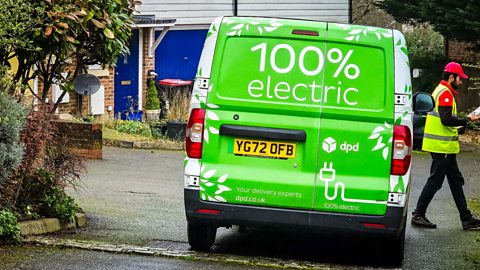 The back of a courier company's delivery van, which states that it's 100% electric - the driver is walking towards a house to deliver a parcel.