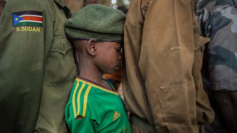 A young boy in a soldier's beret stands between two bigger child soldiers wearing military uniforms tightly squished up against them in a line,