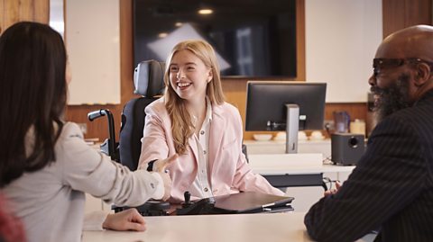 A girl in a wheelchair shaking hands with a female interviewer watched on by a male interviewer.