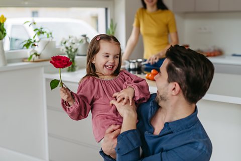 A girl being held by her father in a kitchen holds out a red rose