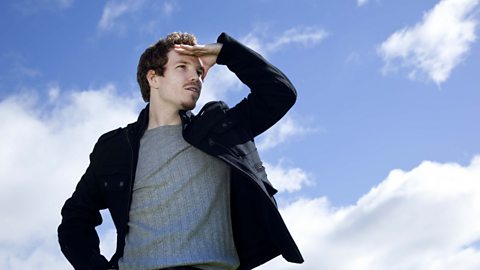 A man holding his hand up to head peering into the distance with a background of a blue sky and clouds behind him.