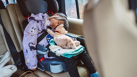 A little girl in a colourful coat sleeps in a car seat whilst holding a teddy bear.