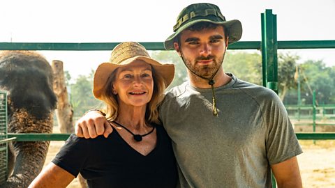 Caroline (left) and Tom are stood in front of an elephant enclosure. Both are wearing sun hats. Caroline is in a black t-shirt and beige hat. Tom is wearing a grey t-shirt and green hat. There is an elephant to the left of the shot.