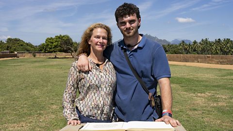 Caroline (left) and Tom, stood in front of the checkpoint book in Kanniyakumari, India. There is grass and trees behind them and blue skies, with wispy clouds. Caroline is wearing a patterned long sleeve shirt. Tom is wearing a blue polo shirt with a crossbody bag