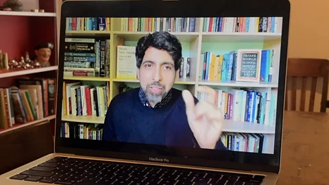BBC Author Sal Khan is shown speaking via videoconference. He can be seen on a laptop monitor wearing a dark blue sweater and gesturing with his hands and bookshelves filled with colourful books are behind him (Credit: BBC)