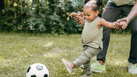 A toddler kicks a football, supported by her parent.