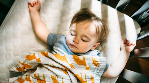 A young child lies sleeping on their back in a cot.