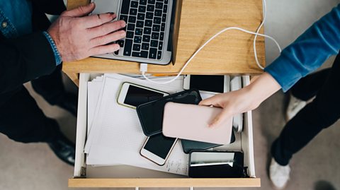 Multiple phones being put inside a desk draw. There is a MACBOOK on top of the desk being worked on by a man.