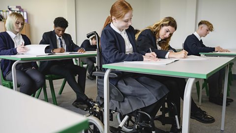 Image of a girl in a wheelchair sitting at a desk in school surrounded by classmates all writing in exam booklets.