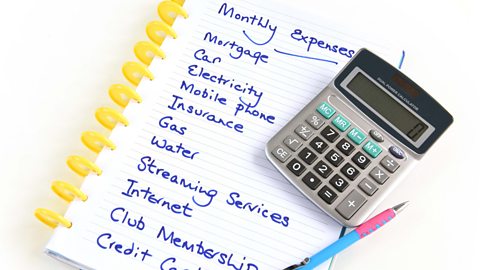 A notepad, pen and calculator with a list of monthly expenses written down in a list - mortgage, car, electricity, mobile phone, insurance, gas, water, streaming services, Internet, club membership and credit card.