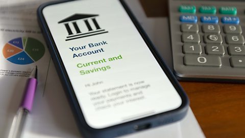 A smart phone sitting on some pages and on the screen it reads 'Your bank account - current and savings - there's also a pen and a calculator beside the phone.