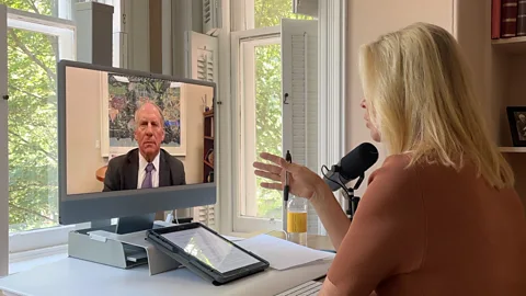 BBC Richard Haass is shown speaking to Katty Kay via videoconference. He can be seen on a computer monitor wearing a black suit, white shirt and purple tie. Kay is shown from behind while wearing a tan-coloured short-sleeved top (Credit: BBC)