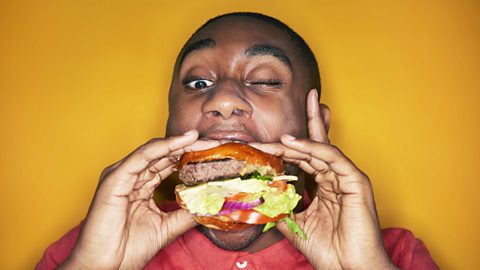 A man eating a burger waering a red shirt and on a yellow backgound.