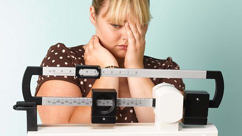 An overweight girl standing on scales and holding her left hand up to her head looking sad - while wearing a brown and white polka dot dress.