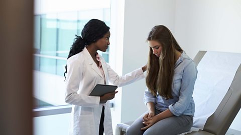 A doctor standing, placing her hand on a depressed looking teenager who is sitting upright.