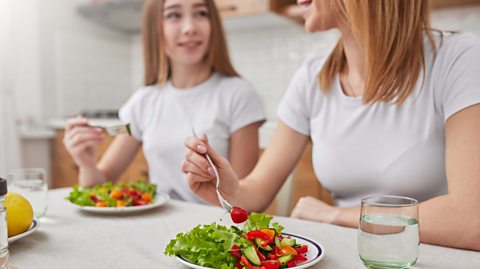 A daughter looking at her mother in the foreground of the image and both are eating salad with glasses of water beside them.