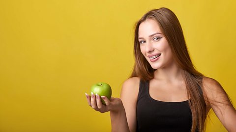 Girl holding an apple in her outstretched hand wearing a black gym top with a yellow background.
