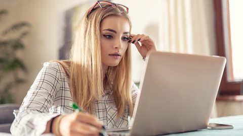 Girl conducting research - sitting at a laptop with a pen - taking notes on what she sees on the screen.